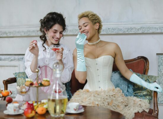 Two people in vintage attire enjoy a tea party with pastries and drinks.