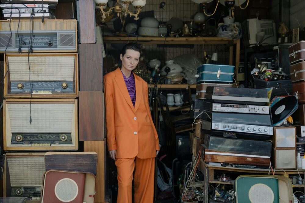Person in orange suit stands amidst vintage electronics and luggage.