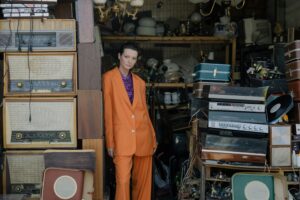 Person in orange suit stands amidst vintage electronics and luggage.