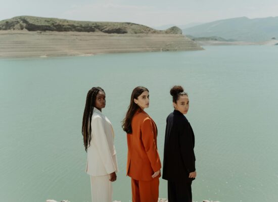 Three people in suits stand by a calm lake with hills.