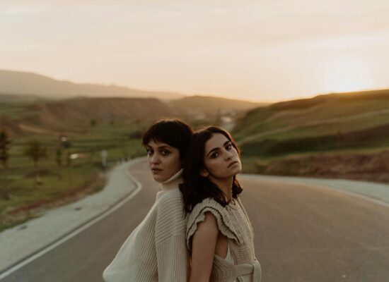 Two people stand back to back on a road at sunset.