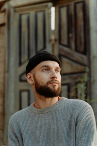 Person wearing a knit sweater and beanie, standing outdoors, thoughtful.