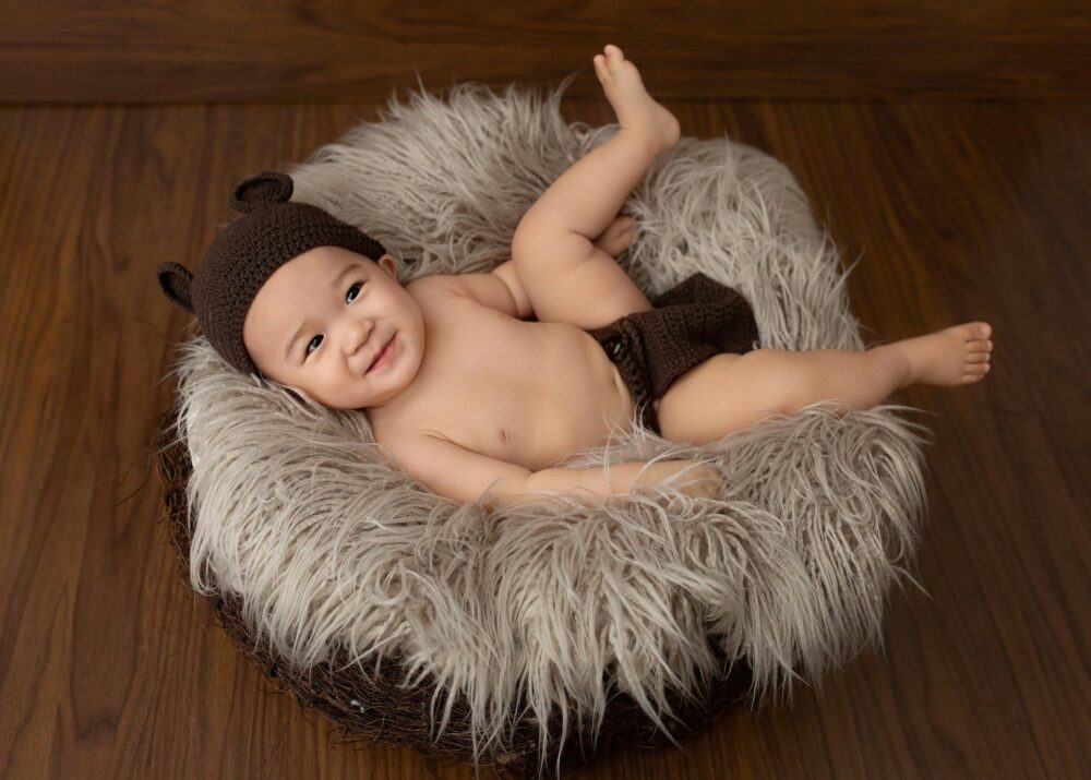 A baby wearing a bear hat and diaper cover lies on a fluffy nest.