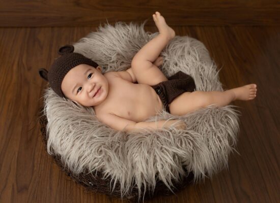 A baby wearing a bear hat and diaper cover lies on a fluffy nest.