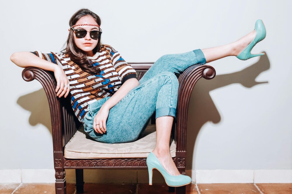 A person sits in a chair wearing jeans, a patterned shirt, and teal heels.