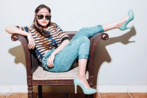 A person sits in a chair wearing jeans, a patterned shirt, and teal heels.
