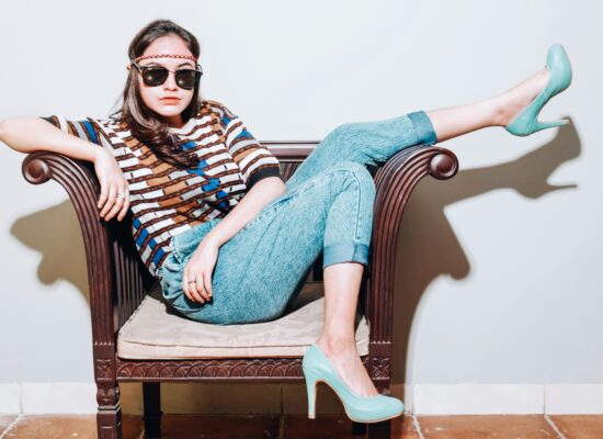 A person sits in a chair wearing jeans, a patterned shirt, and teal heels.