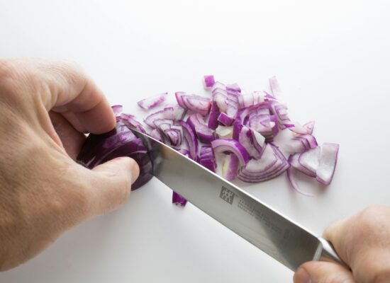 Hands chopping a red onion with a knife on white surface.