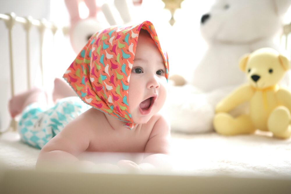 A baby wearing a colorful bandana lies on its stomach with toys.