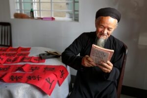 An elderly person reads a book next to red calligraphy scrolls.
