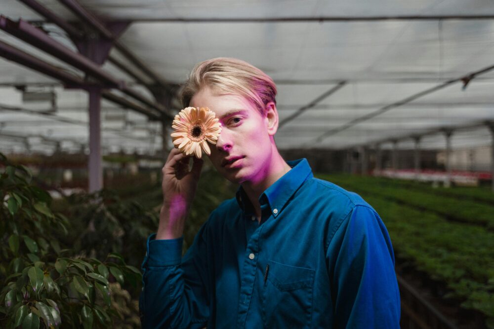 Person holding a flower over one eye in a greenhouse.