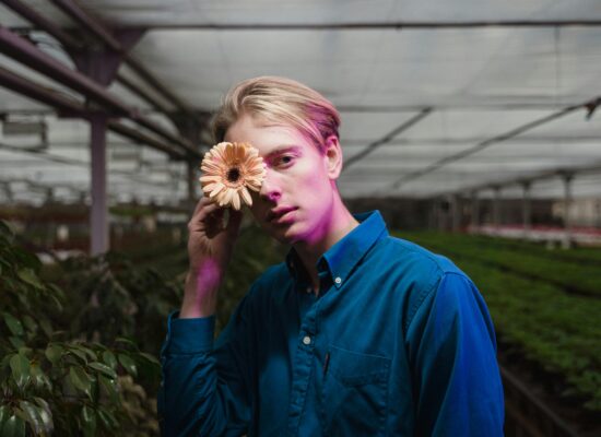 Person holding a flower over one eye in a greenhouse.