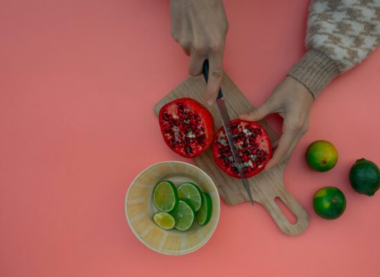 Hands cutting pomegranate, limes in bowl, pink background, wooden board.
