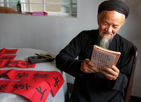 An elderly person reads a book next to red calligraphy scrolls.