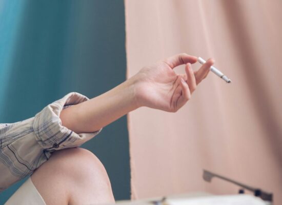 Person holding cigarette, sitting by typewriter, pastel background curtains.