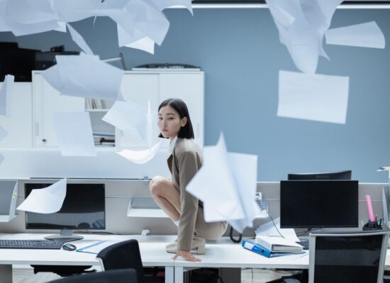 Person crouching on office desk, papers flying everywhere, chaotic scene.