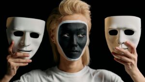 Person with painted face holds two white masks, black background.