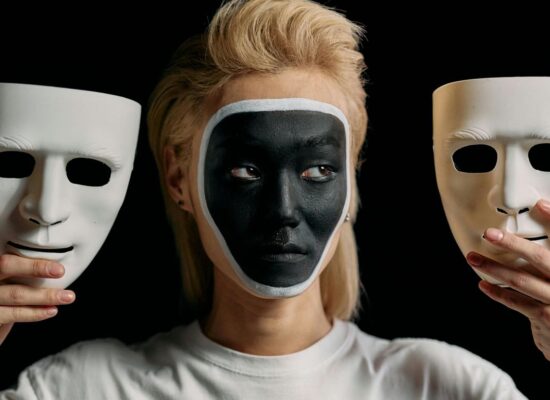 Person with painted face holds two white masks, black background.