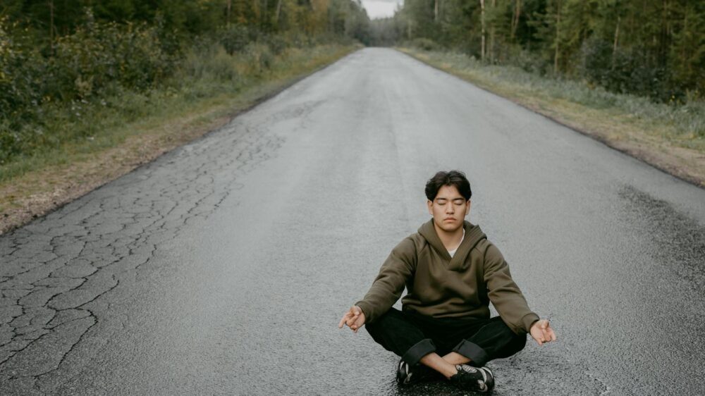 Person meditating on empty road surrounded by green forest.