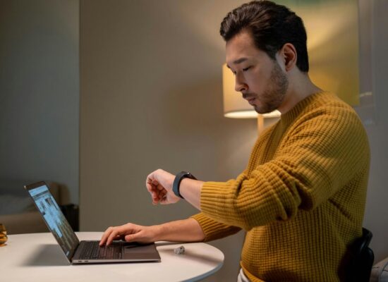 Person at desk using laptop, checking smartwatch, cozy indoor setting.