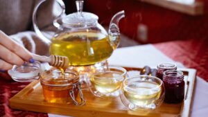 Glass teapot, tea cups, honey, and jam on wooden tray.
