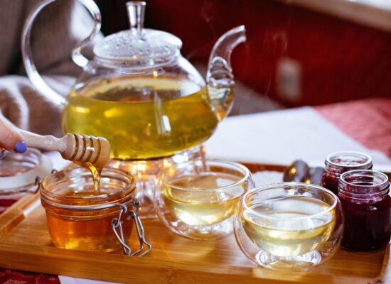 Glass teapot, tea cups, honey, and jam on wooden tray.