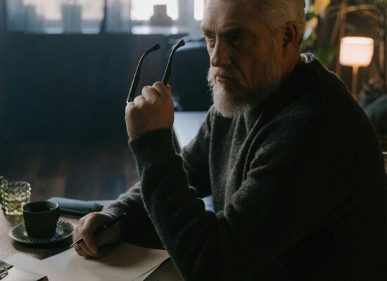 Person sitting at desk, holding glasses, thoughtful, cozy room.