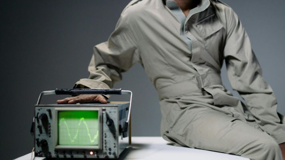 Person in jumpsuit with hand on oscilloscope displaying waveform.