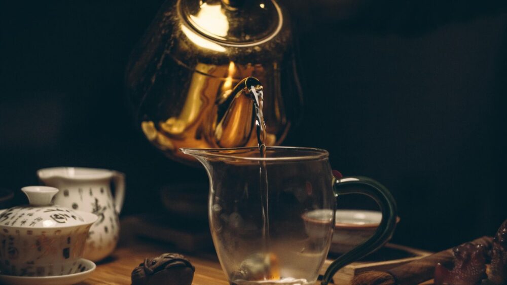 Golden teapot pouring water into a glass pitcher during tea ceremony.