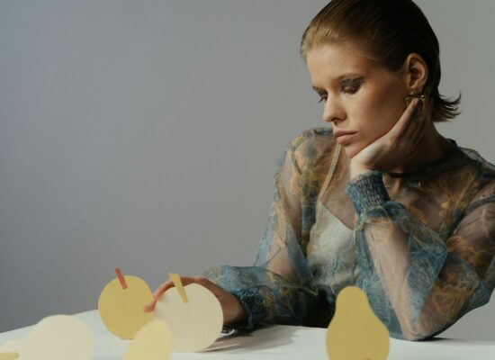 Person with hand on chin near paper fruit shapes on table.