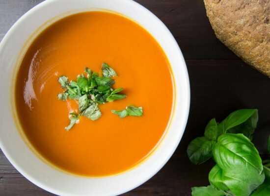 Bowl of tomato soup with herbs, bread, and basil leaves.