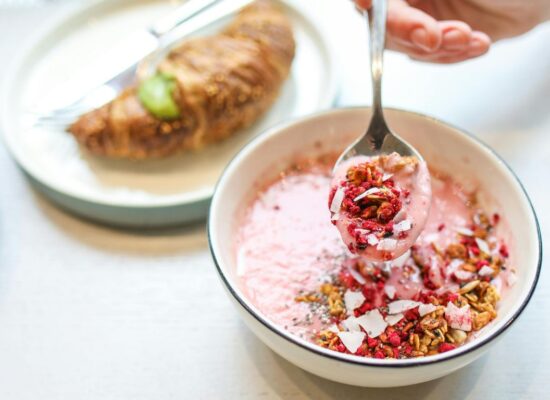 Bowl of pink yogurt with granola, croissant in background.