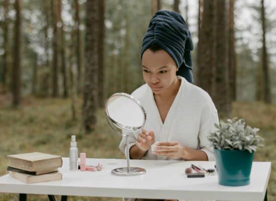 Person sitting at table in forest, applying skincare, mirror nearby.