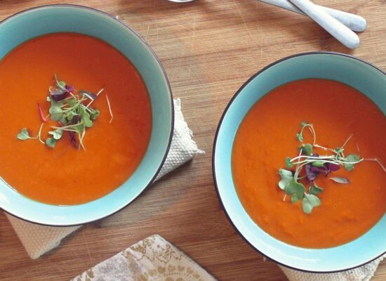 Two bowls of tomato soup with garnish, bread, and spoons.