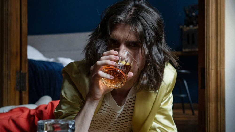 Person lying on floor, drinking whiskey, decanter in foreground.