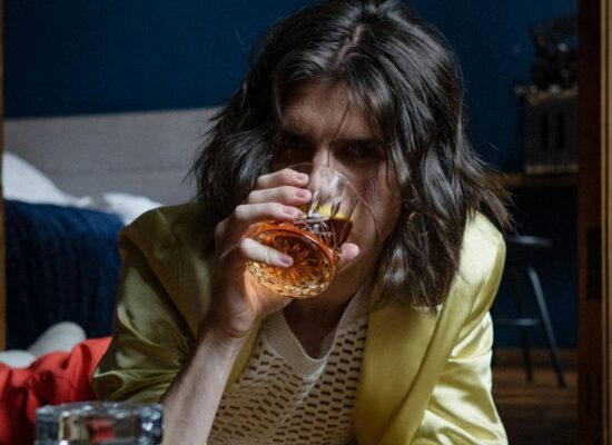 Person lying on floor, drinking whiskey, decanter in foreground.