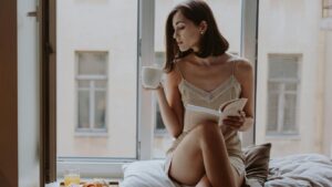 Person sitting by window, drinking coffee, reading book, breakfast nearby.