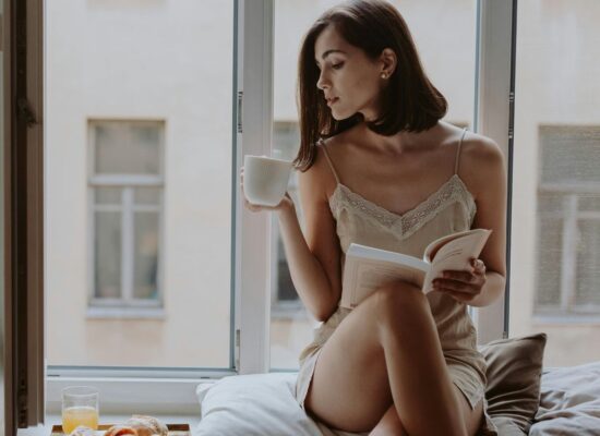 Person sitting by window, drinking coffee, reading book, breakfast nearby.