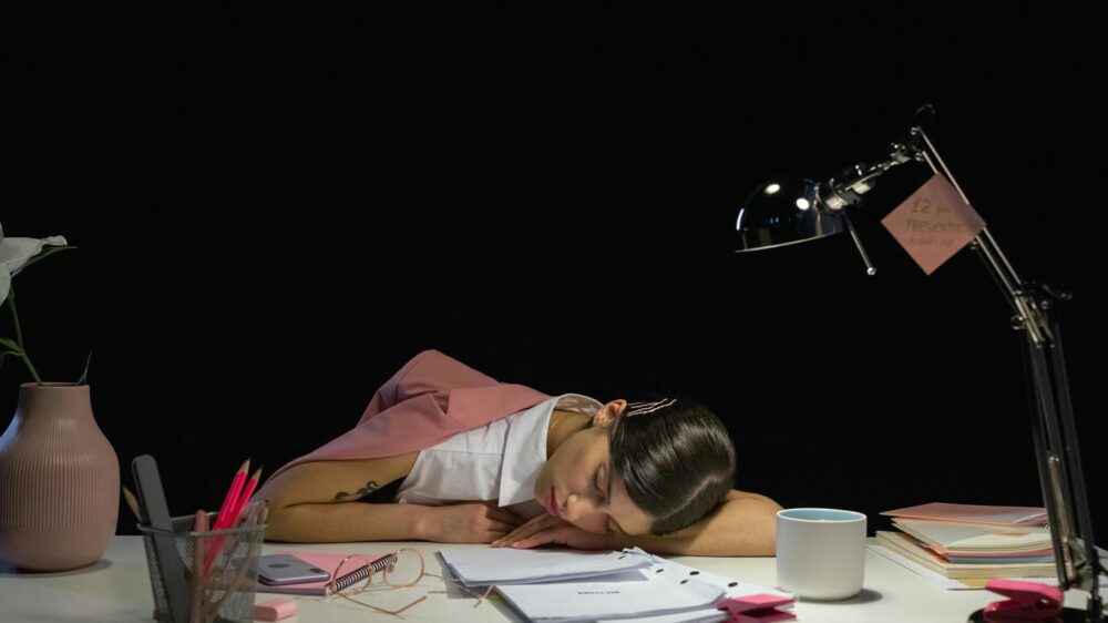 Person sleeping at a desk with papers and a lamp.
